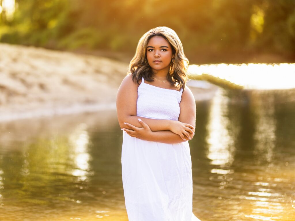 Senior portraits of a high school senior in a white dress by a river in the sunset during summer in Ridgefield Washington