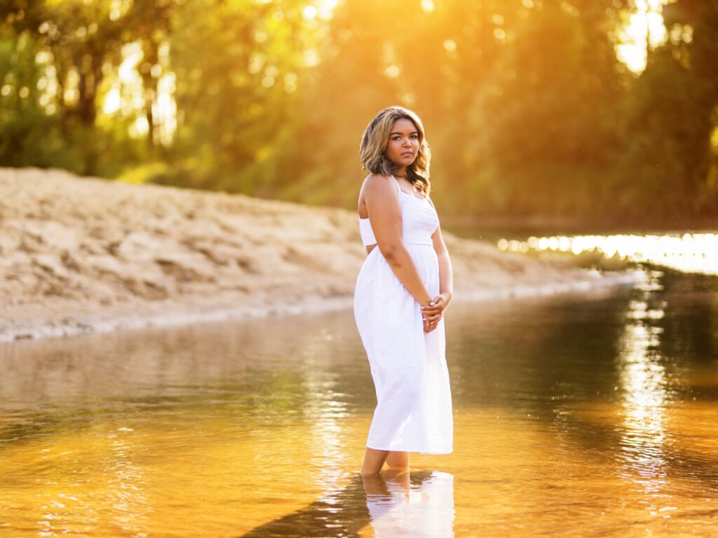 Senior portraits of a high school senior in a white dress by a river in the sunset during summer in Ridgefield Washington