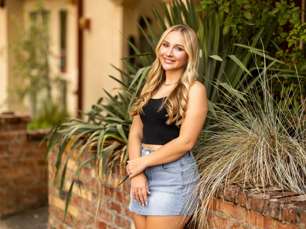 Girl senior photos in Lake Oswego, OR in the late summer during golden hour and she's wearing a simple black top with jean skirt in a city setting with bricks and palm fronds which give a Mediterranean look to the photo.