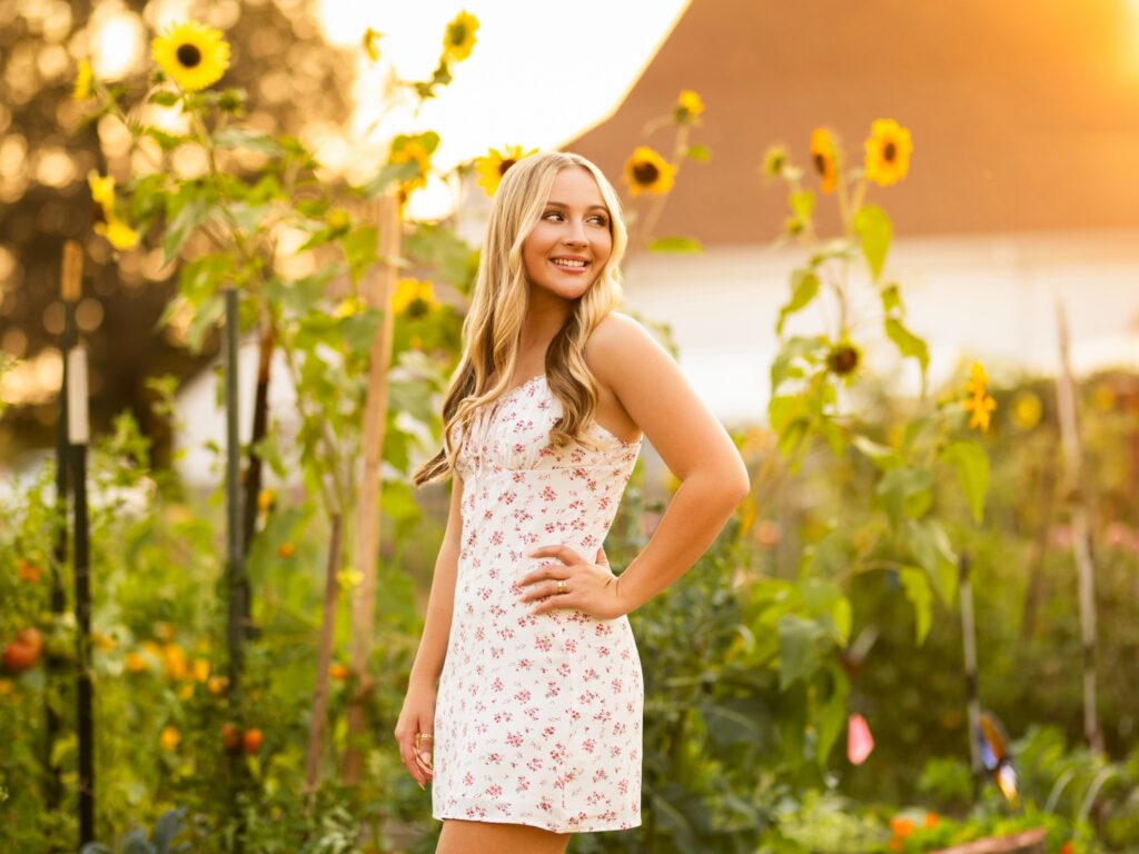 Girl senior photos in Lake Oswego, OR in the late summer during golden hour and she's wearing a floral dress in a field of sunflowers with a white barn in the background and the sun behind her
