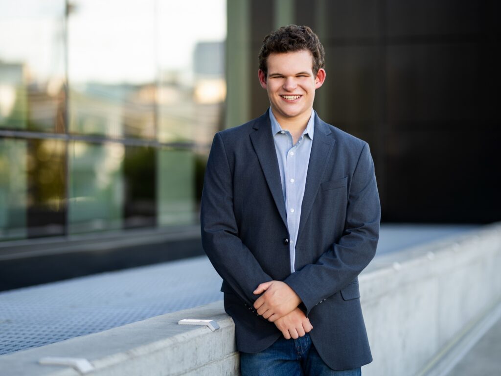 Guy senior photos in downtown Portland, OR wearing a gray sport coat, light blue button-up shirt, and jeans with modern glass architecture.