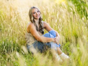 Girl senior pictures in Vancouver, WA during summer wearing a floral cami and distressed jeans while sitting in tall golden grass with soft afternoon sunlight.