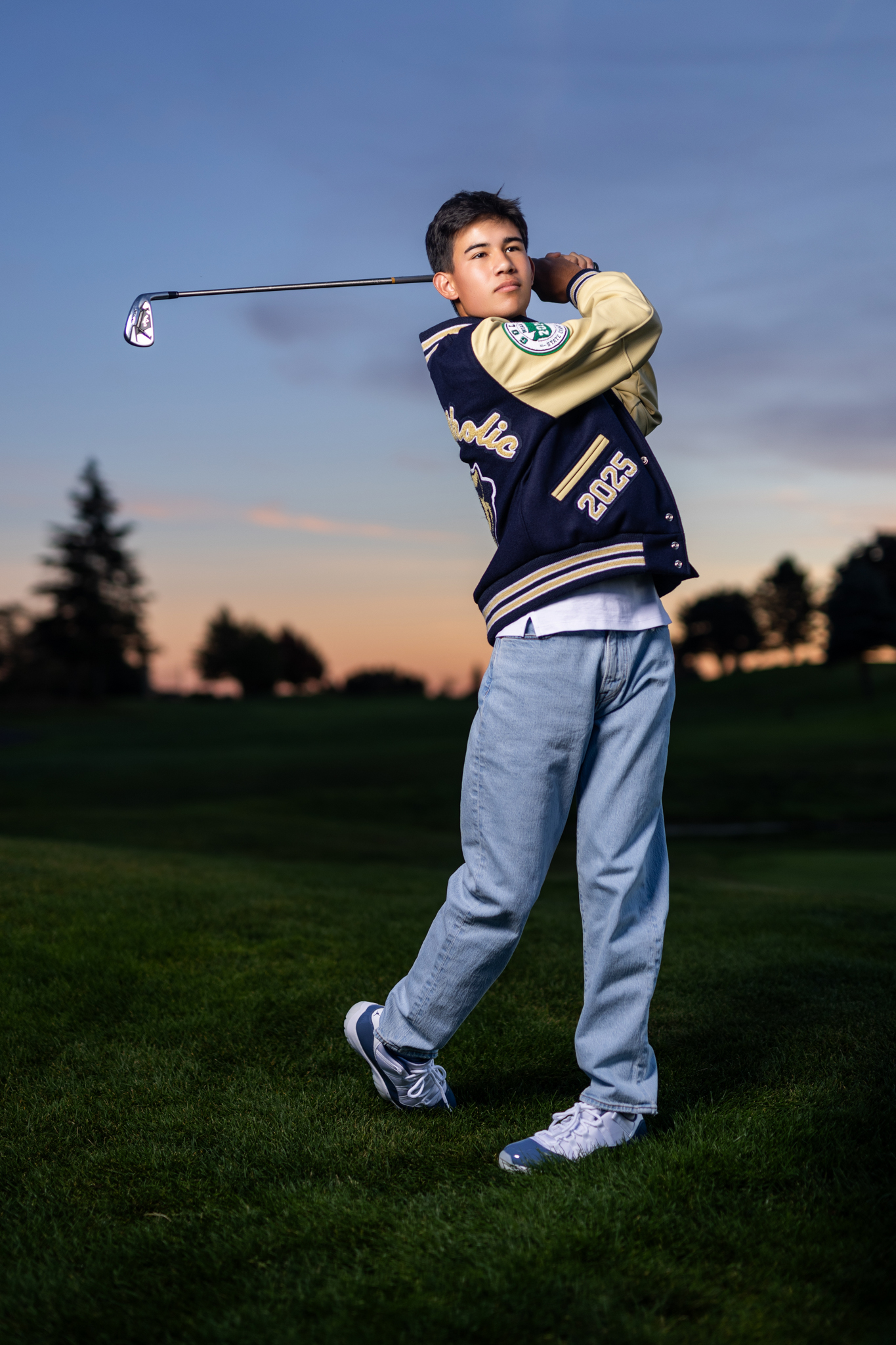 Guy senior photos on a golf course in Vancouver, WA during sunset wearing a letterman jacket and jeans while posing with a golf club.