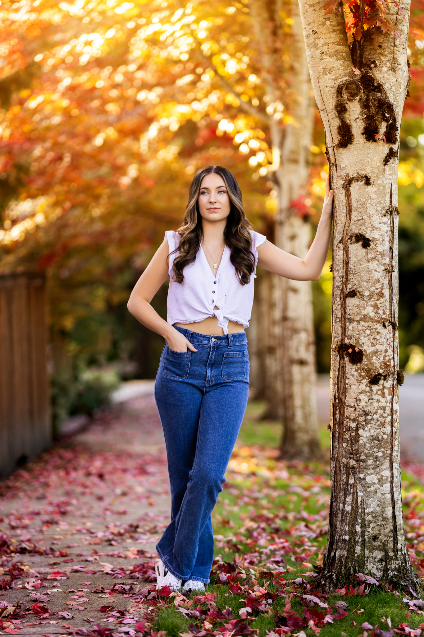 Girl senior photos in Vancouver, WA during the fall wearing a white tie-front top and jeans on a sidewalk covered with colorful autumn leaves.