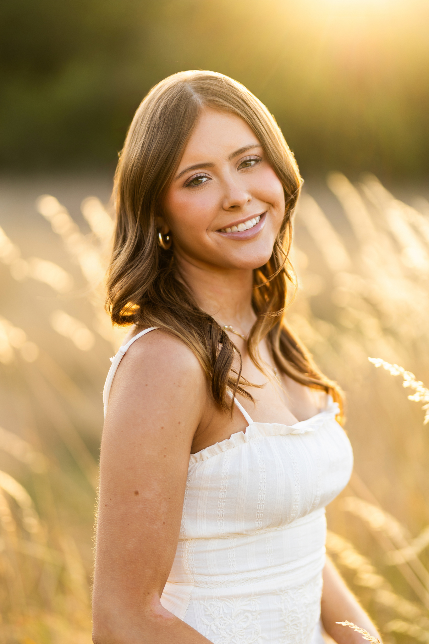 Girl senior photos in a tall grass field during golden hour wearing a white dress with warm sunlight behind her.